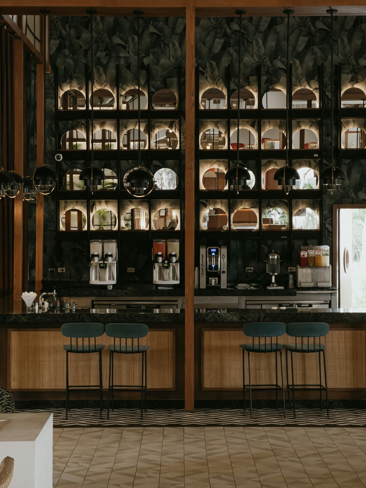 A hotel bar with lit shelves, black pendant lamps and four stools at a rattan-fronted counter.
