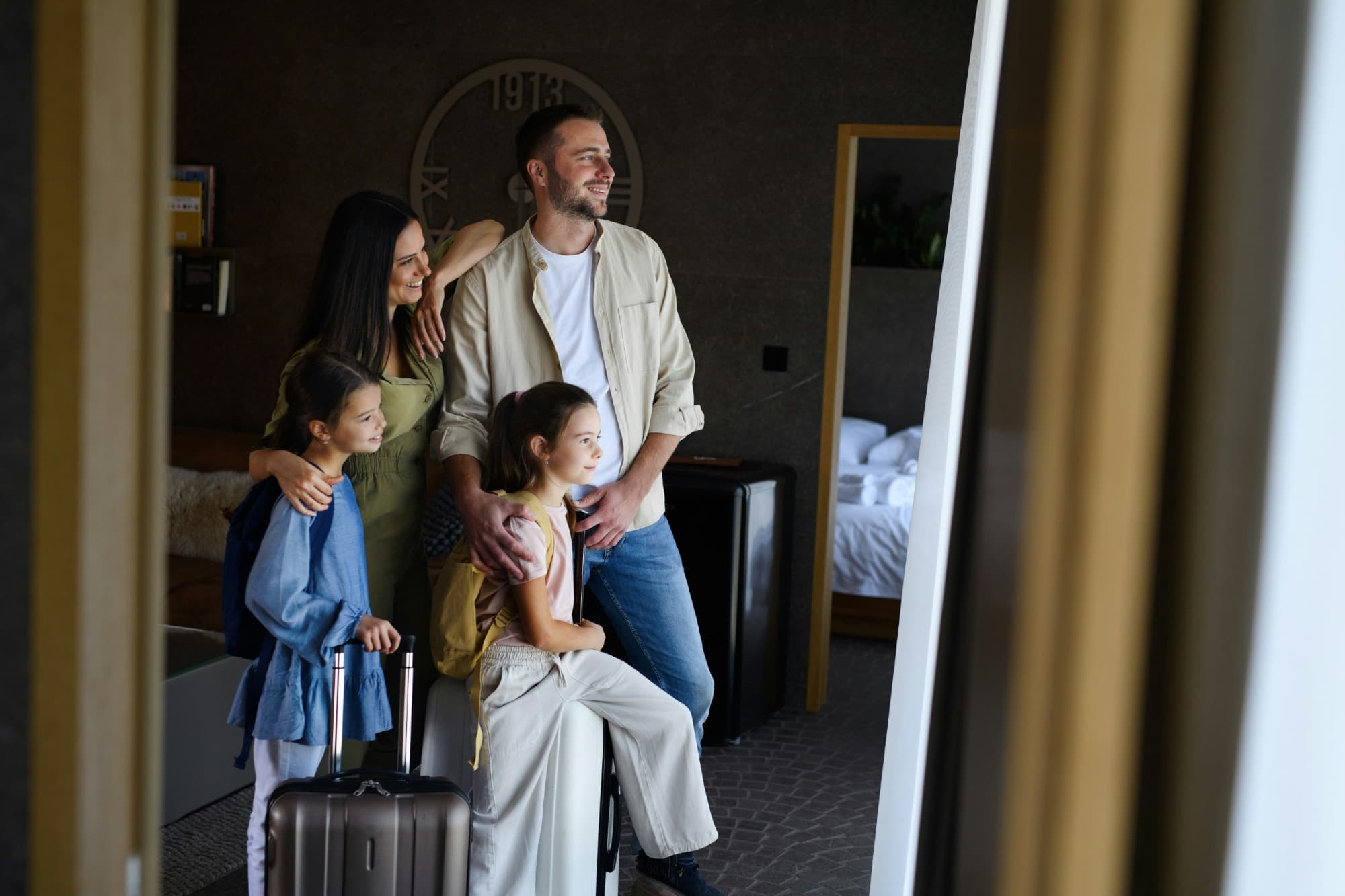 A family of four with suitcases and backpacks, smiling as they look into their hotel room.