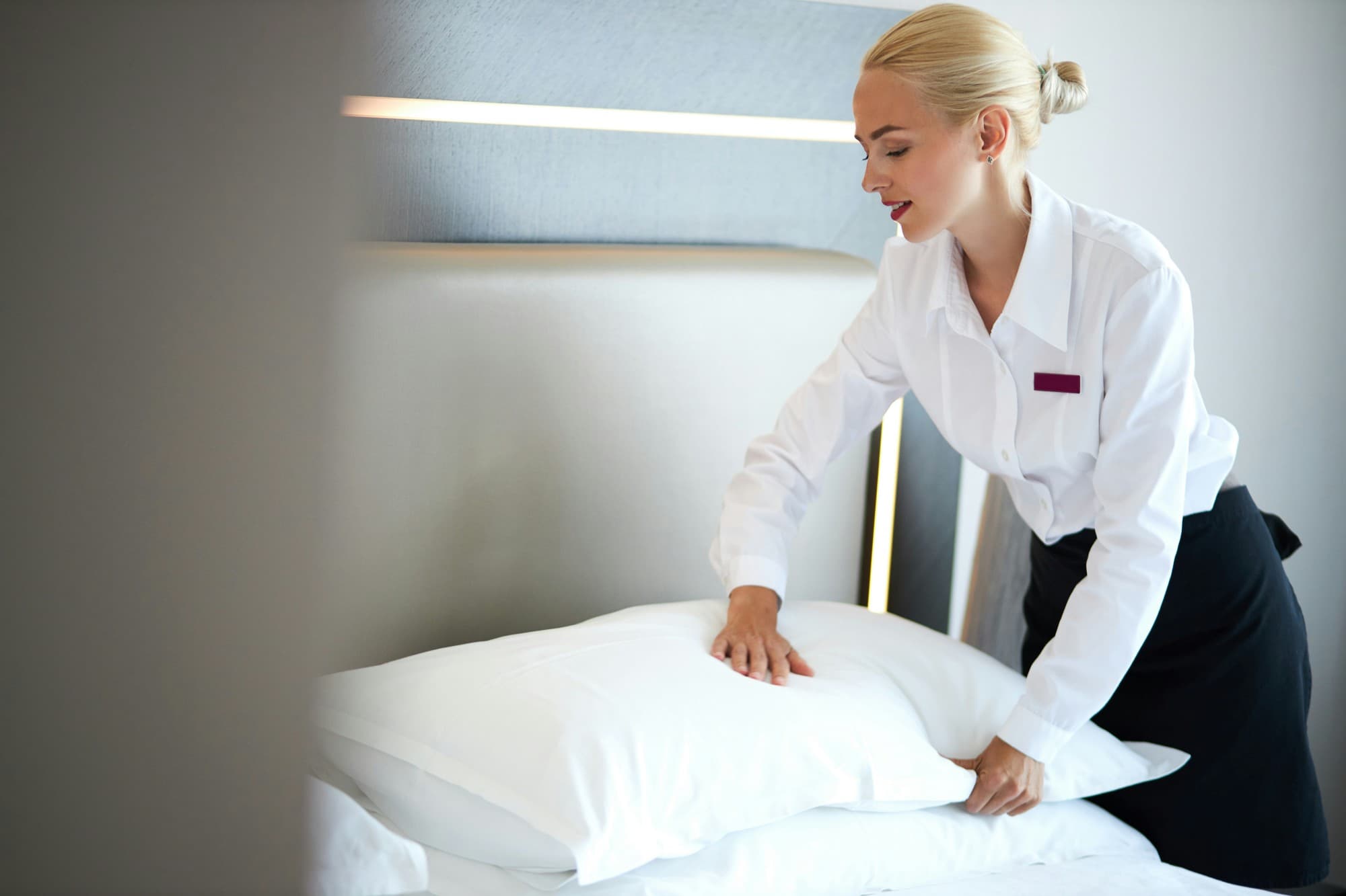 A housekeeper in a white shirt plumping the pillows on a freshly made hotel bed.