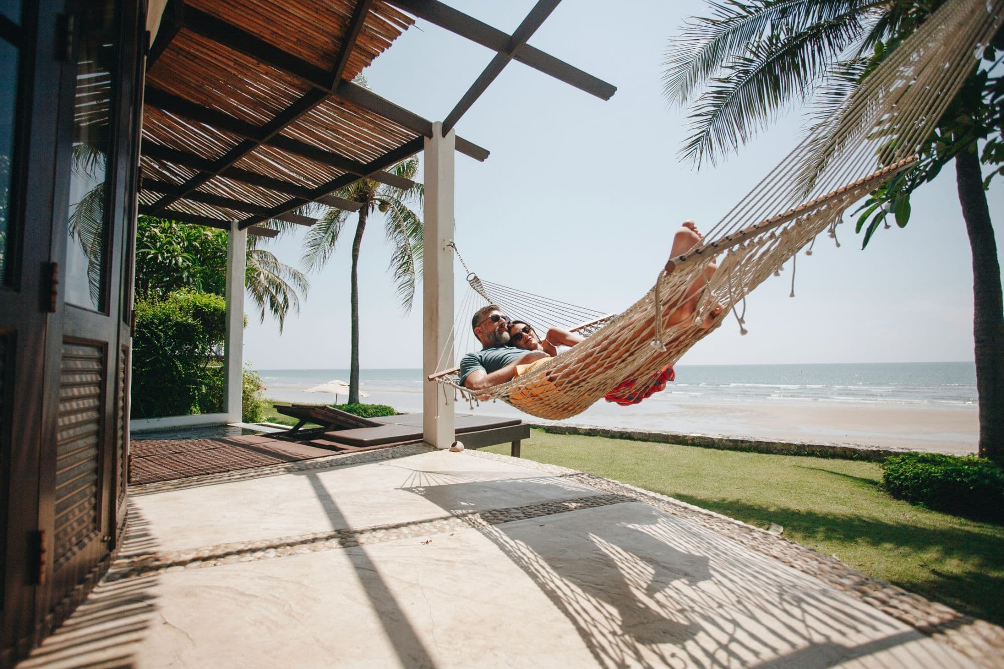 Two guests relaxing together in a hammock on the terrace of a beachfront rental, the sea behind them.