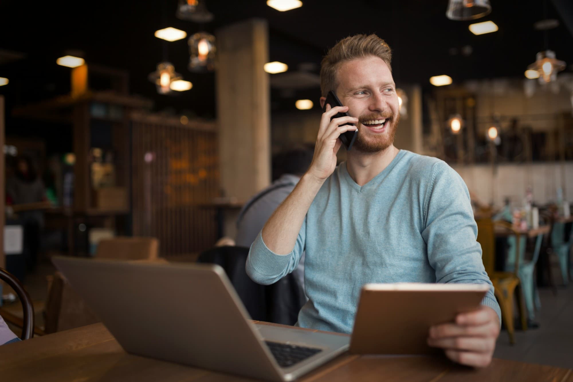 A smiling homeowner on a phone call at a café table, a tablet in his other hand and a laptop open in front of him.